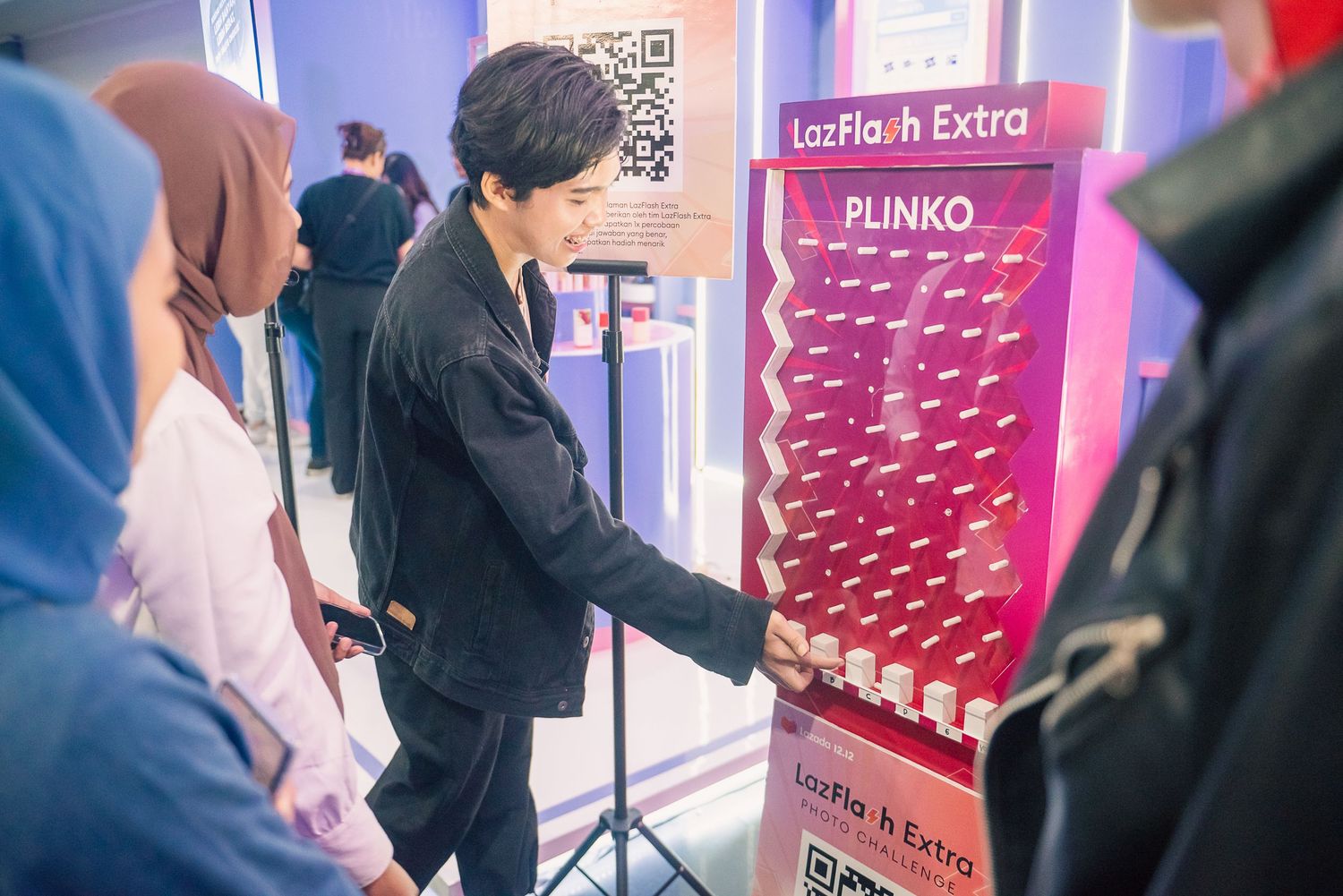 Young man playing Plinko game at LazFlash Extra promotional event booth with onlookers watching nearby.