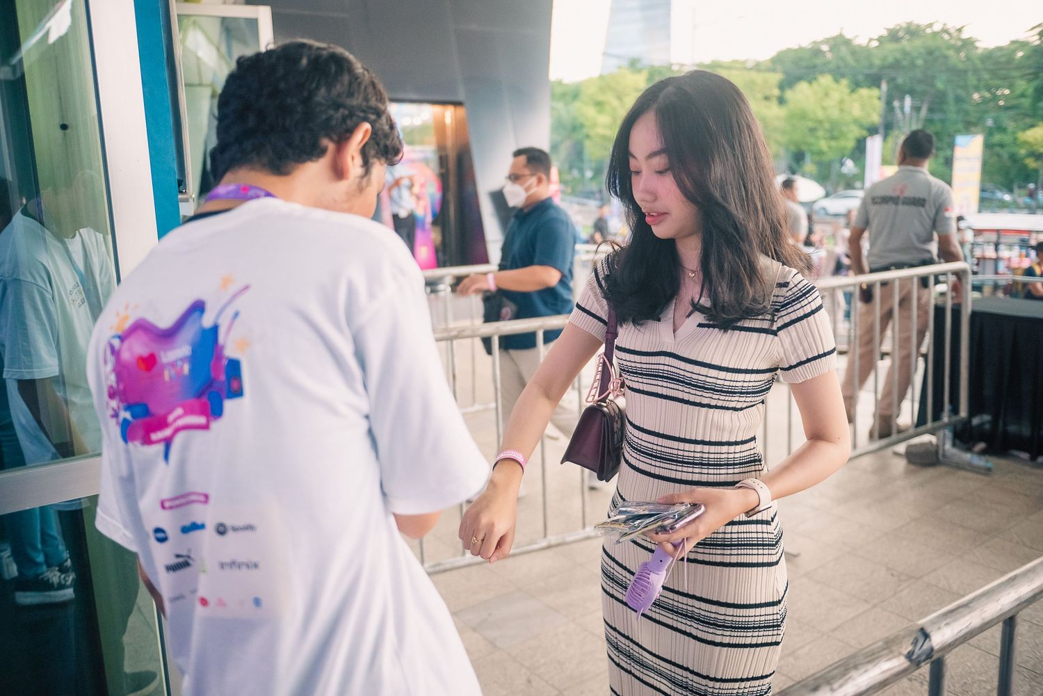 A woman in a striped dress shows her ticket to a staff member in a white t-shirt at an outdoor event entrance.
