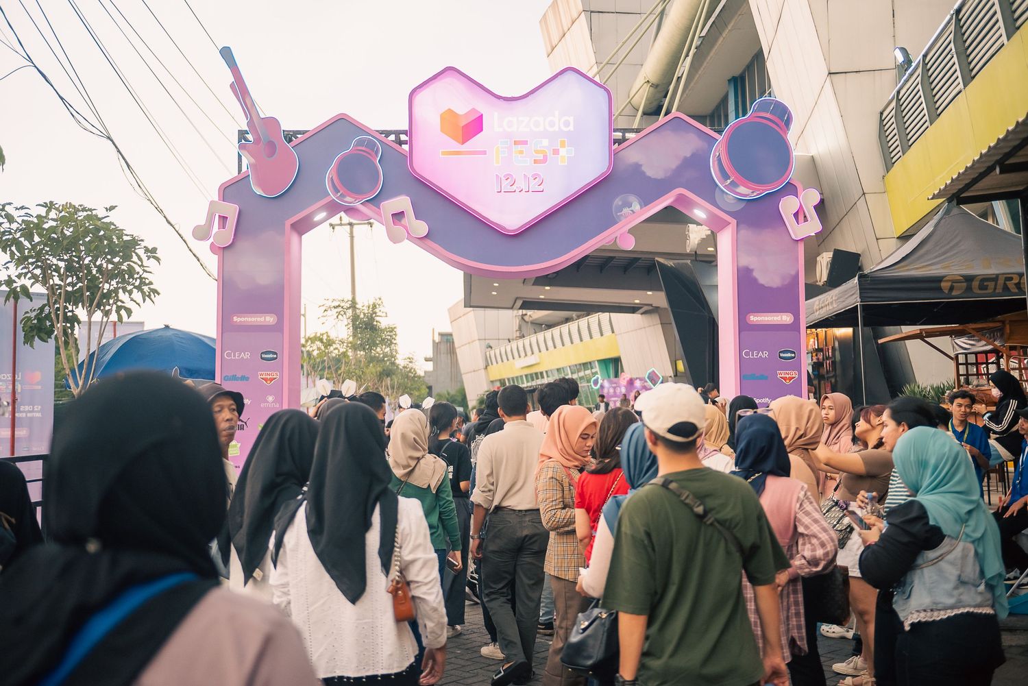 Crowd of people gathered under a pink decorative arch at an outdoor street festival event.