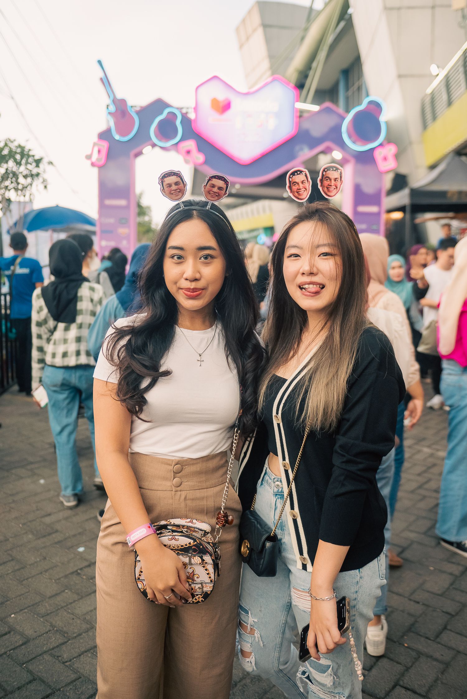 Two young women smiling at a colorful neon festival entrance with a crowd gathered in the background.