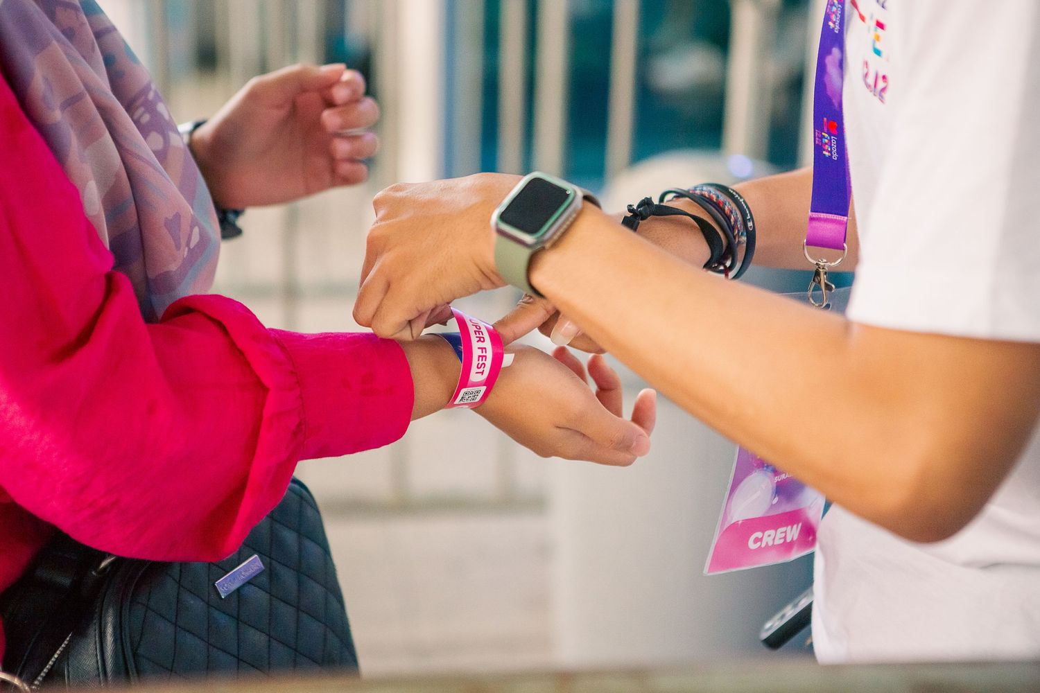 A crew member attaches a pink wristband to an attendee's wrist at an event, both wearing smartwatches.