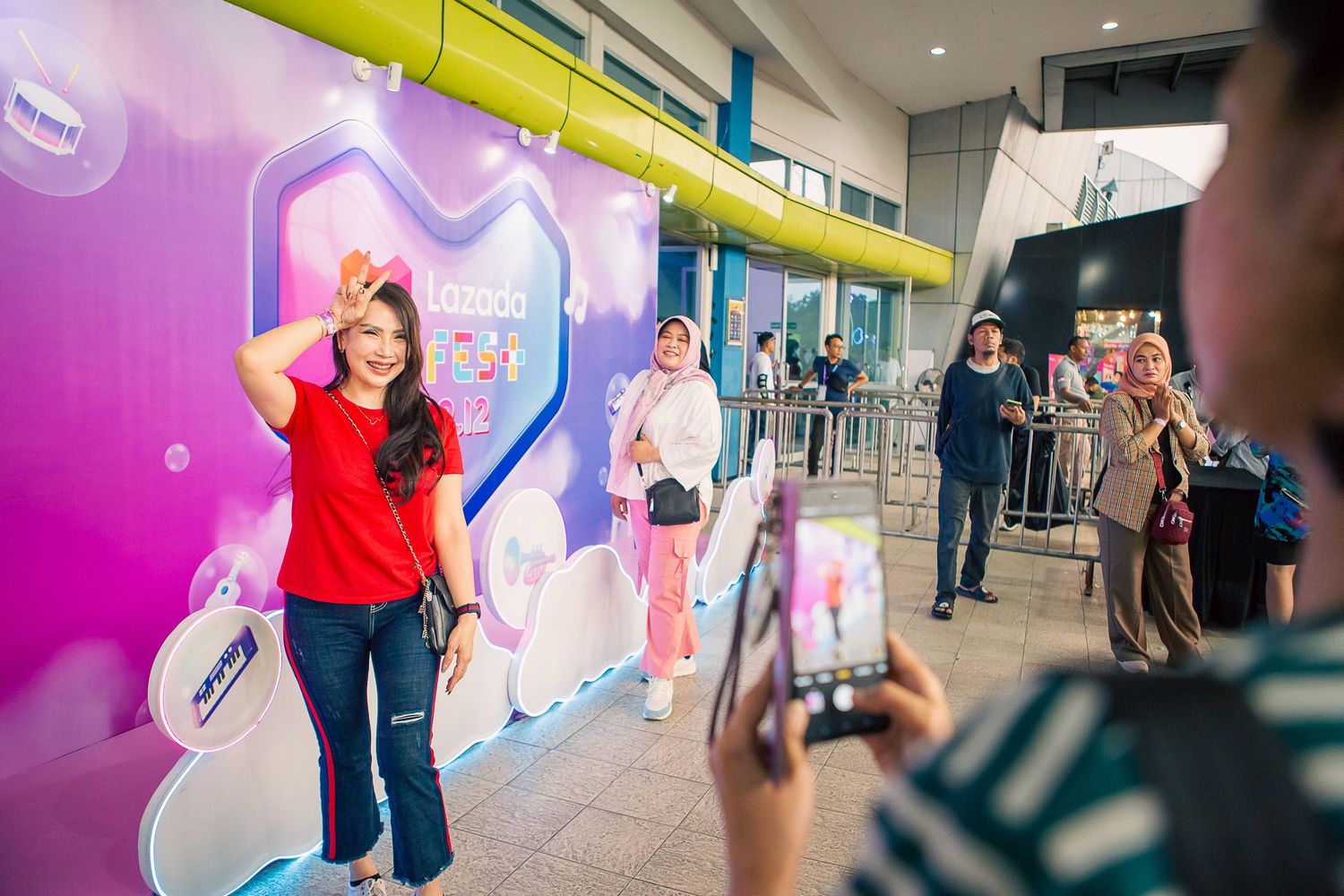 Woman in red shirt poses for photo at colorful promotional event backdrop while others watch nearby.