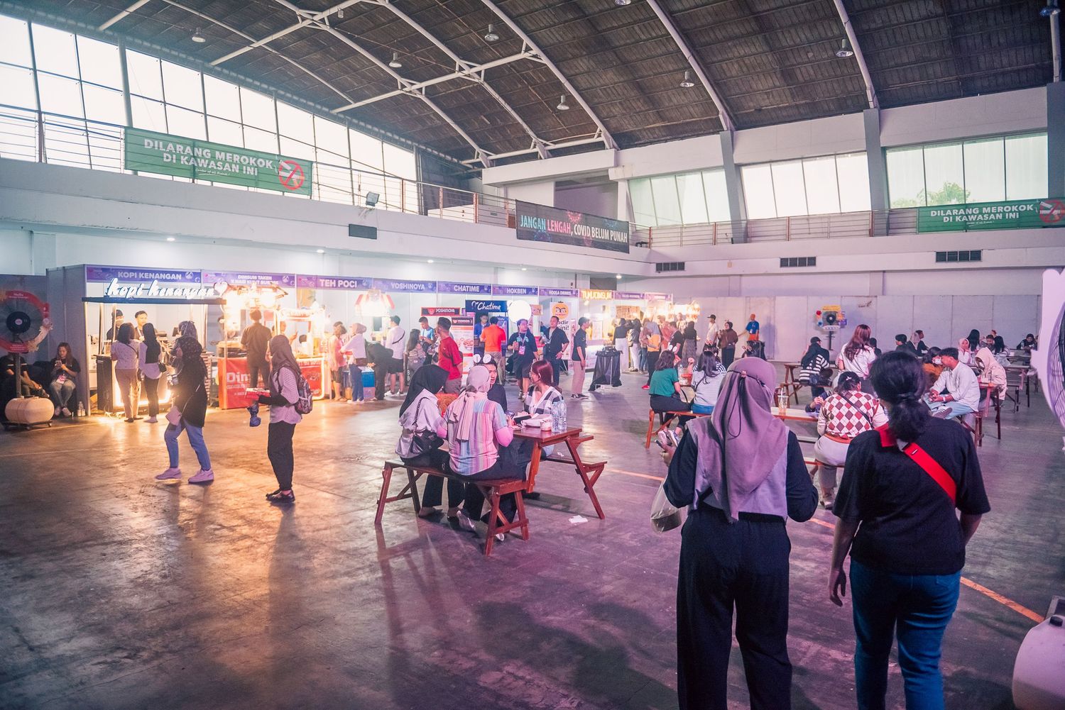 Crowds of people browse vendor booths and exhibits inside a large indoor event hall with colorful lighting.