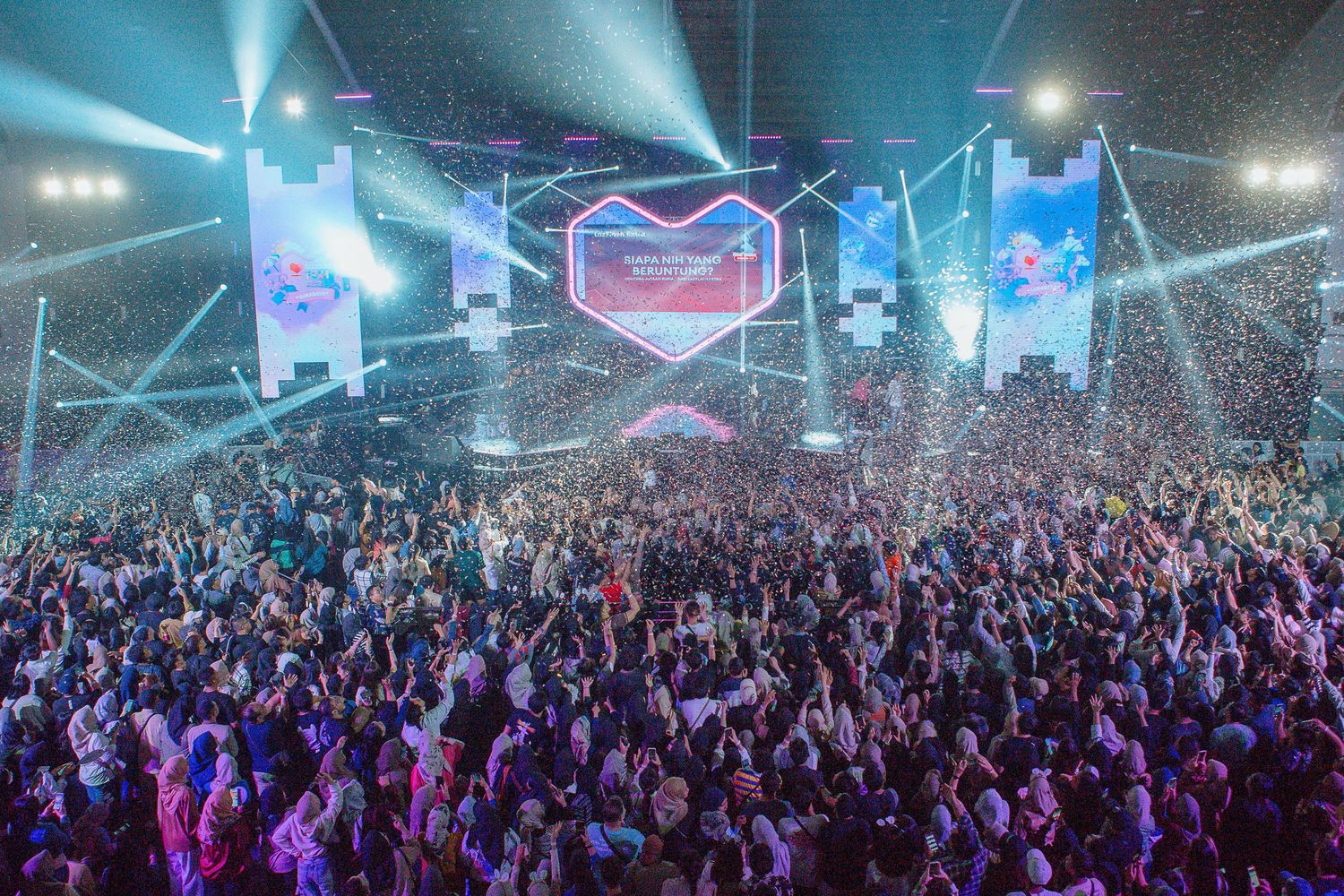 Massive concert crowd cheers under colorful stage lights and confetti at a vibrant music festival event.