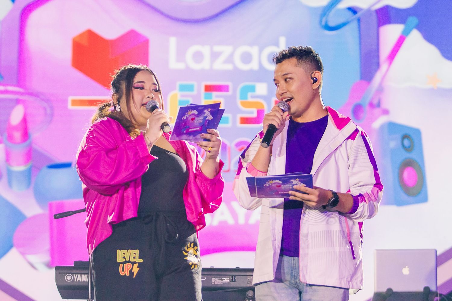 Two hosts present on stage at a colorful Lazada Festival event, holding microphones and tablets under bright lights.
