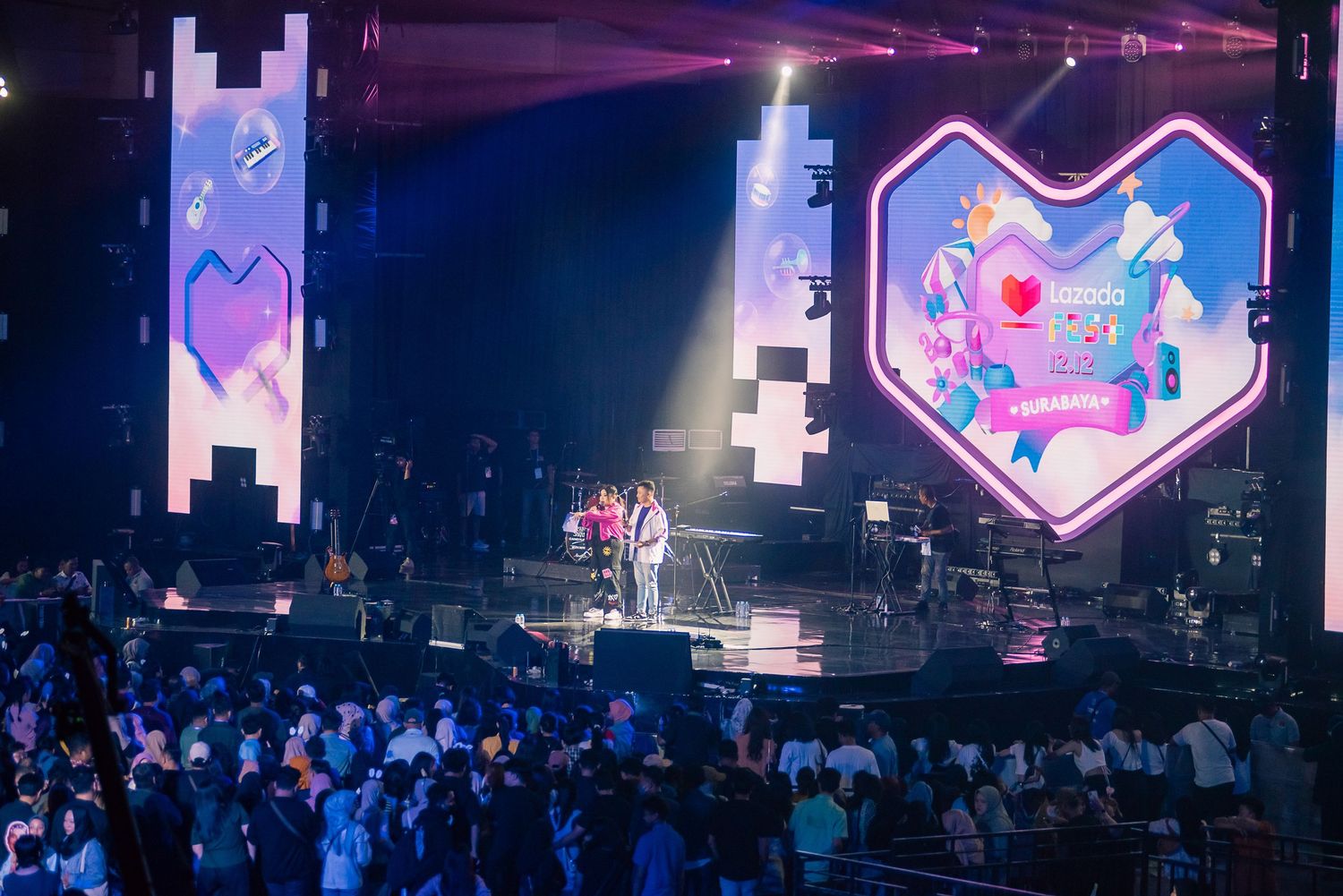 Live concert stage with colorful LED displays, heart-shaped screen, and large crowd at an indoor music event.