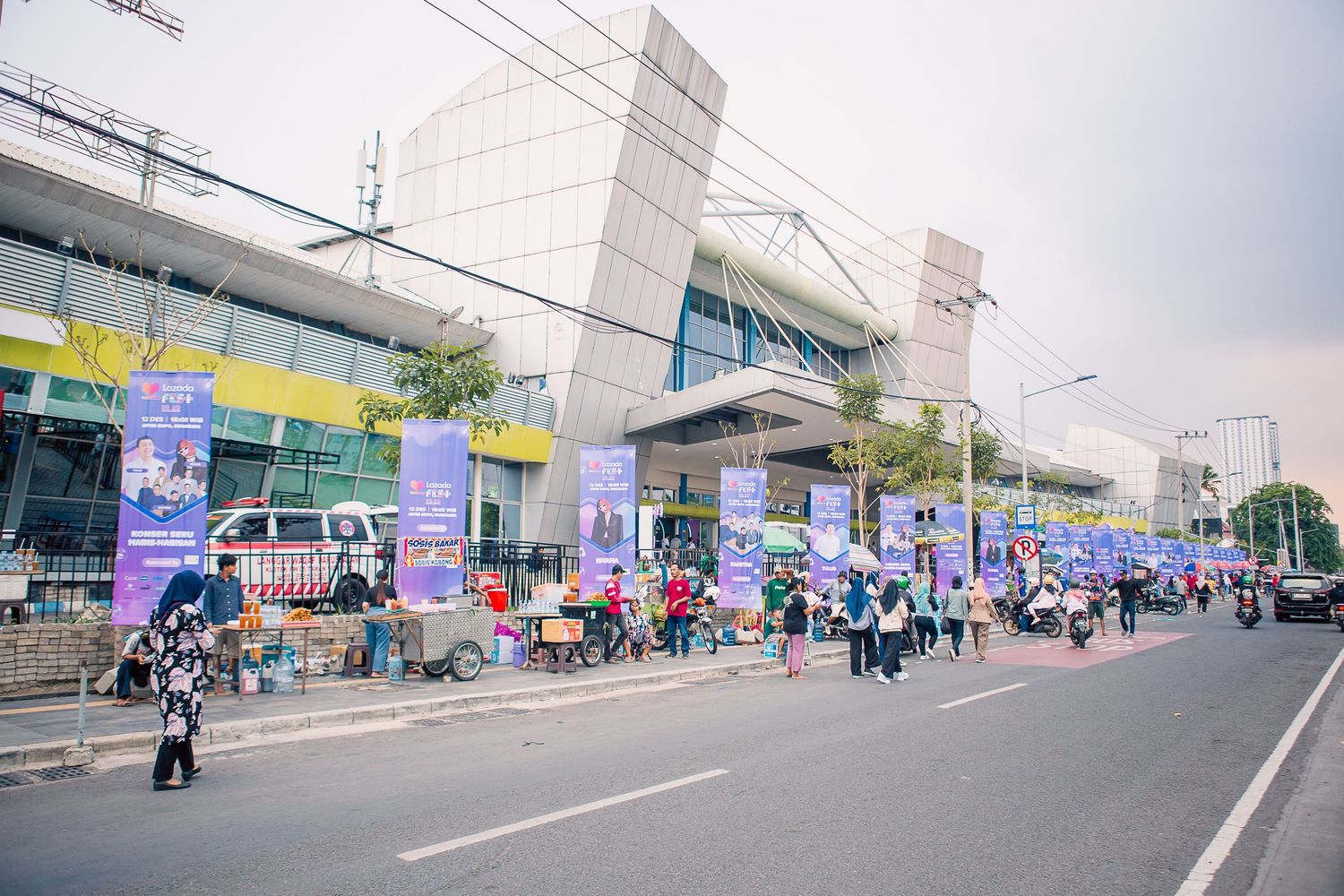 Busy street scene outside a modern shopping mall with colorful banners and crowds of people on the sidewalk.