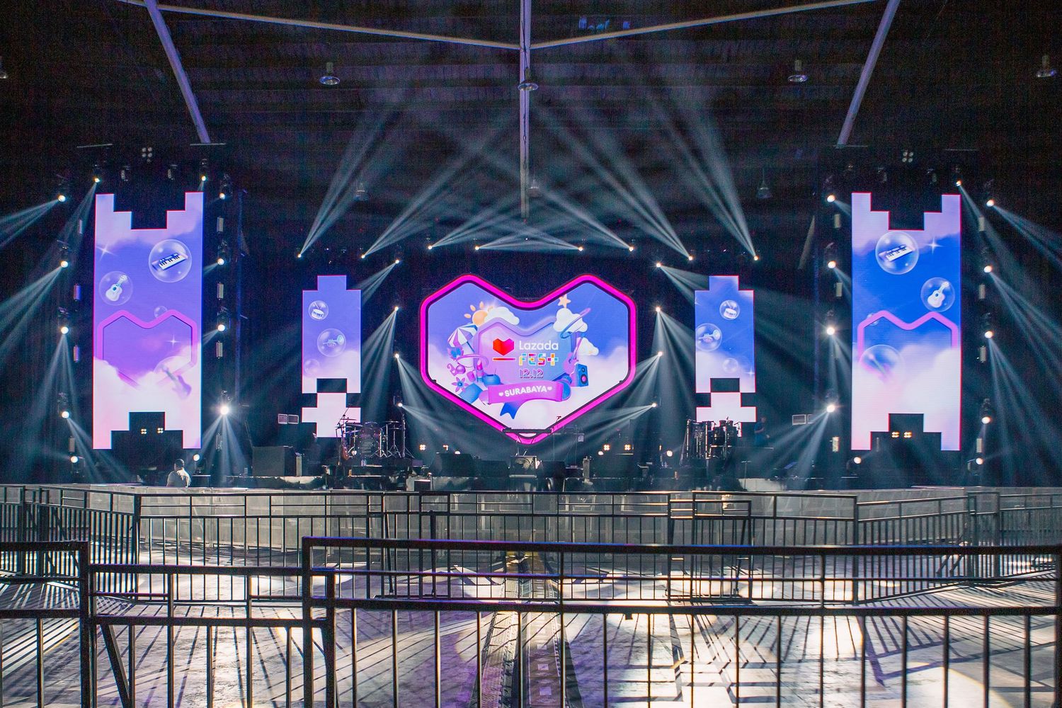 Large concert stage with vibrant LED screens displaying colorful heart graphic, dramatic light beams in an empty arena.