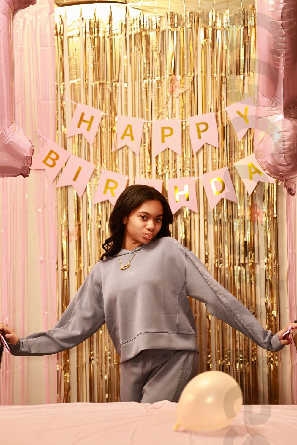 Young woman in gray sweatsuit poses at birthday party with gold tinsel backdrop and Happy Birthday banner.