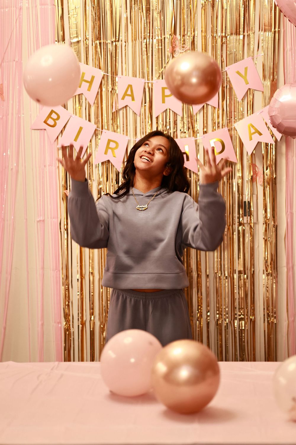 Joyful teen tossing pink gold balloons at 13th birthday party with glittery gold fringe backdrop.