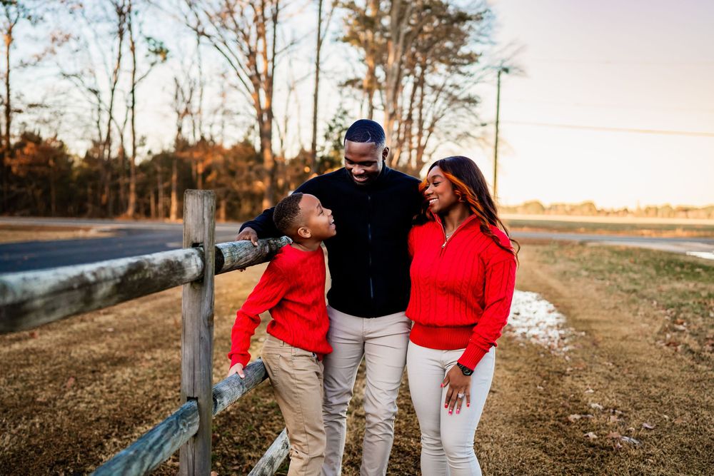 Happy Black family of three smiling together outdoors by a wooden fence in matching red sweaters.
