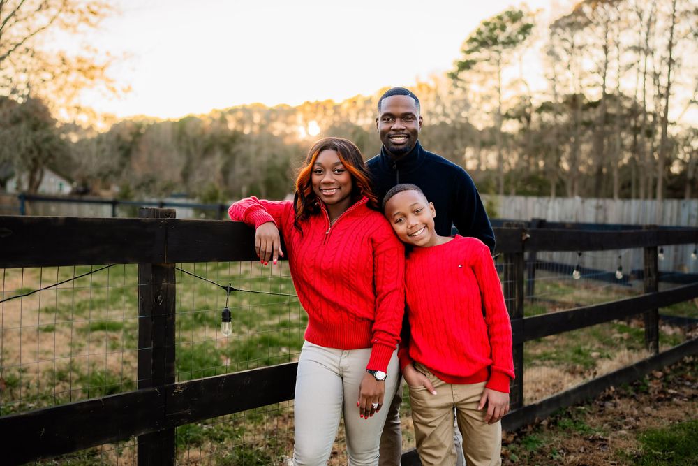 A joyful Black family of three wearing red sweaters poses by a wooden fence in a sunny outdoor setting.