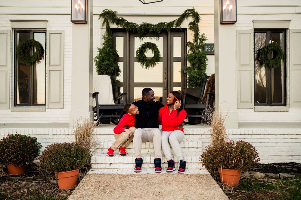 Family of three in red and black outfits sitting on white porch steps decorated with Christmas wreaths and garland.