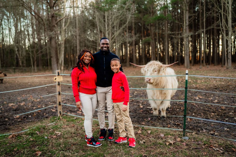 Family of three posing with a fluffy white goat at a farm, wearing matching red outfits among trees.
