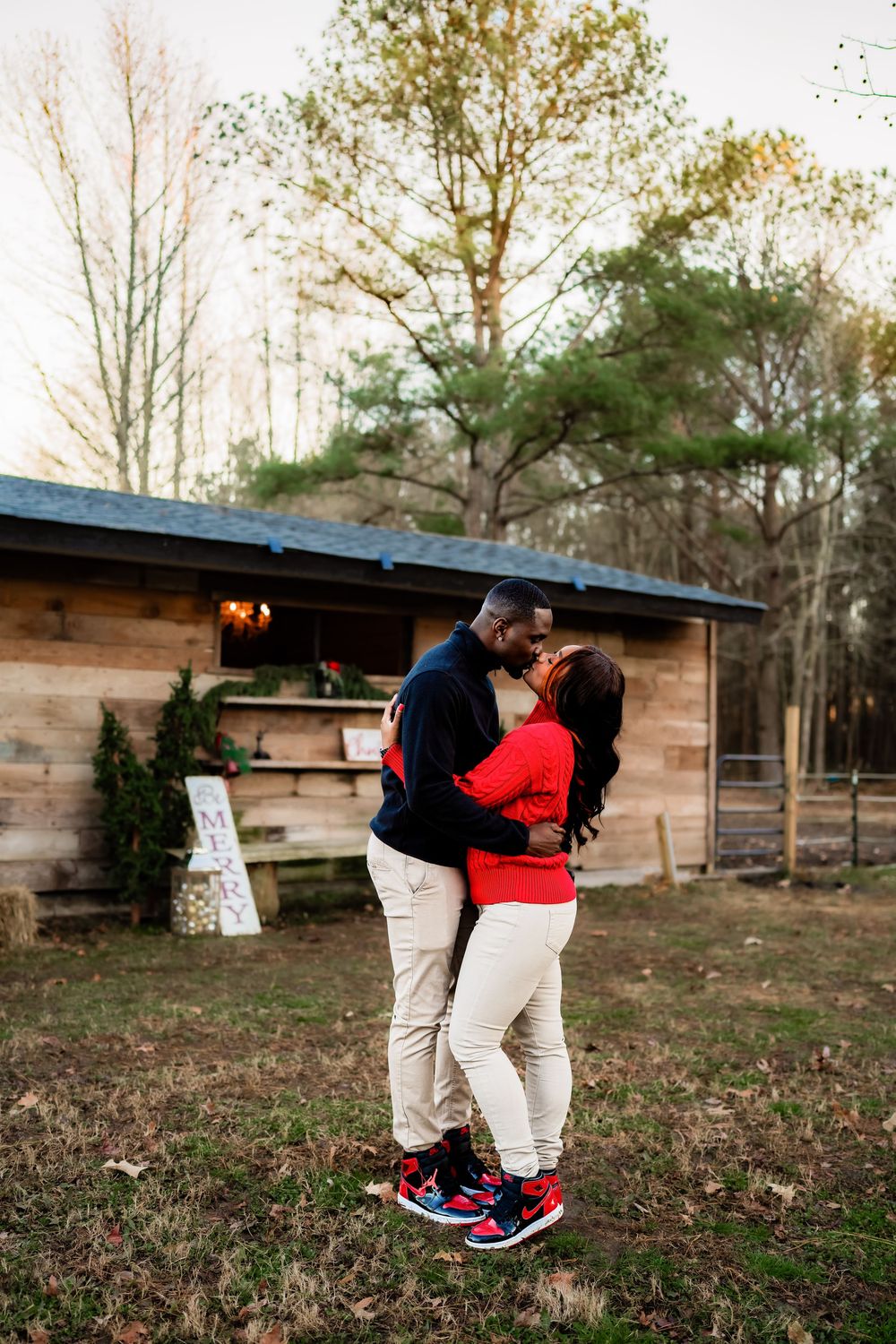 Couple sharing a romantic kiss outdoors near a rustic wooden cabin surrounded by trees at sunset.