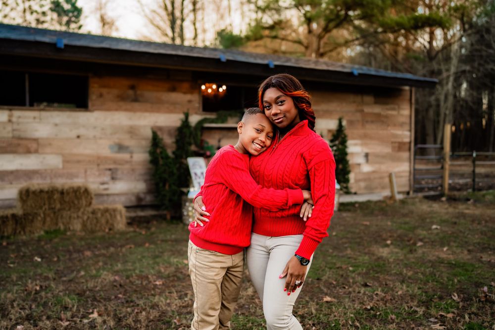 Mother and child sharing a warm embrace outdoors, both wearing matching red sweaters near a rustic barn.