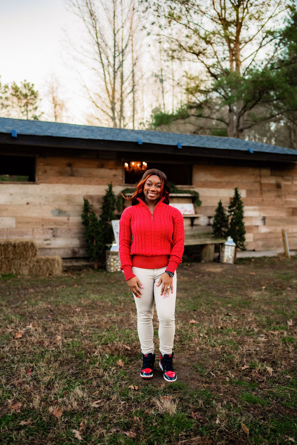 A smiling woman in a red sweater and white pants stands outdoors in front of a rustic wooden building surrounded by trees.