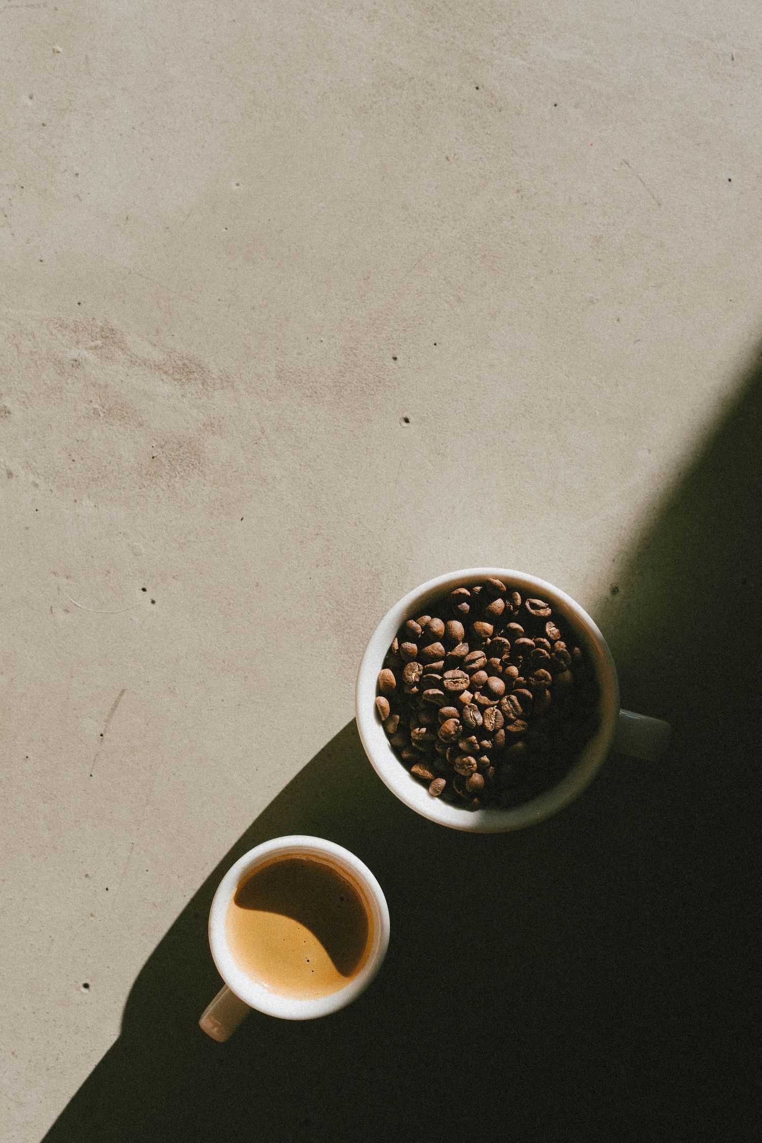 Overhead view of two white ceramic cups, one filled with roasted coffee beans and one with espresso, on a beige surface.