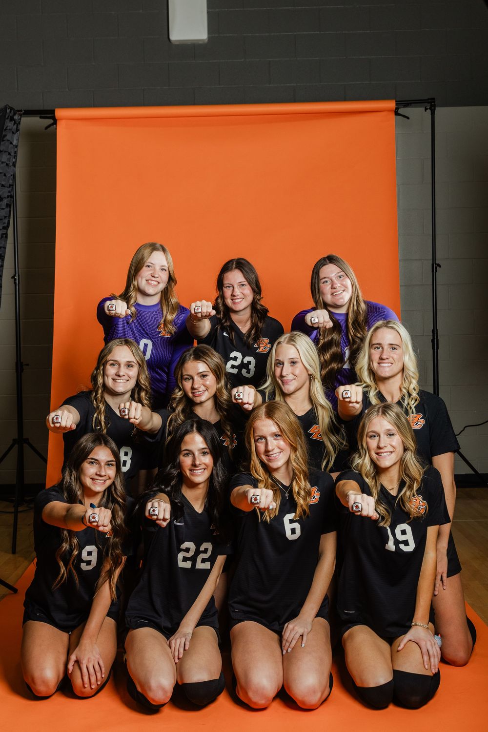 Volleyball team in black uniforms kneels together for team photo against orange and dark background.
