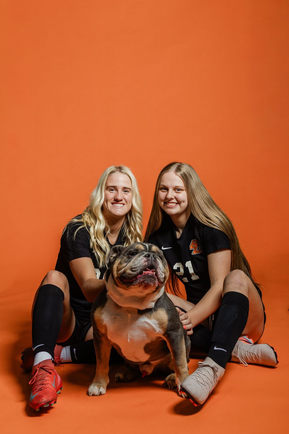 Two athletes pose playfully with a dog on orange studio background.