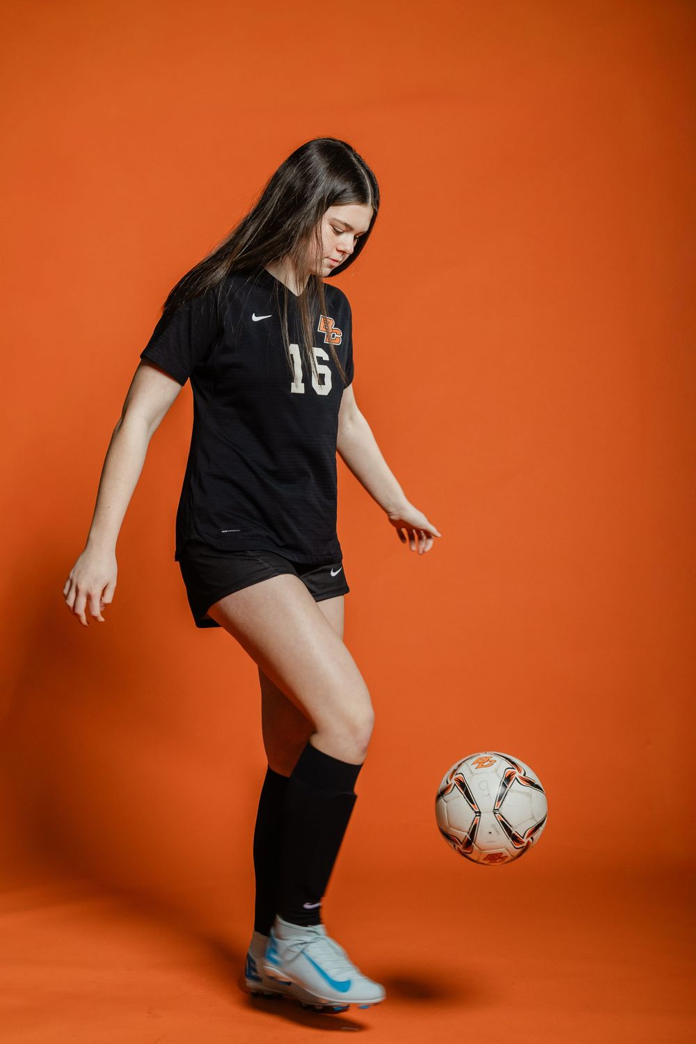 A soccer player in black uniform practices ball control against an orange backdrop.