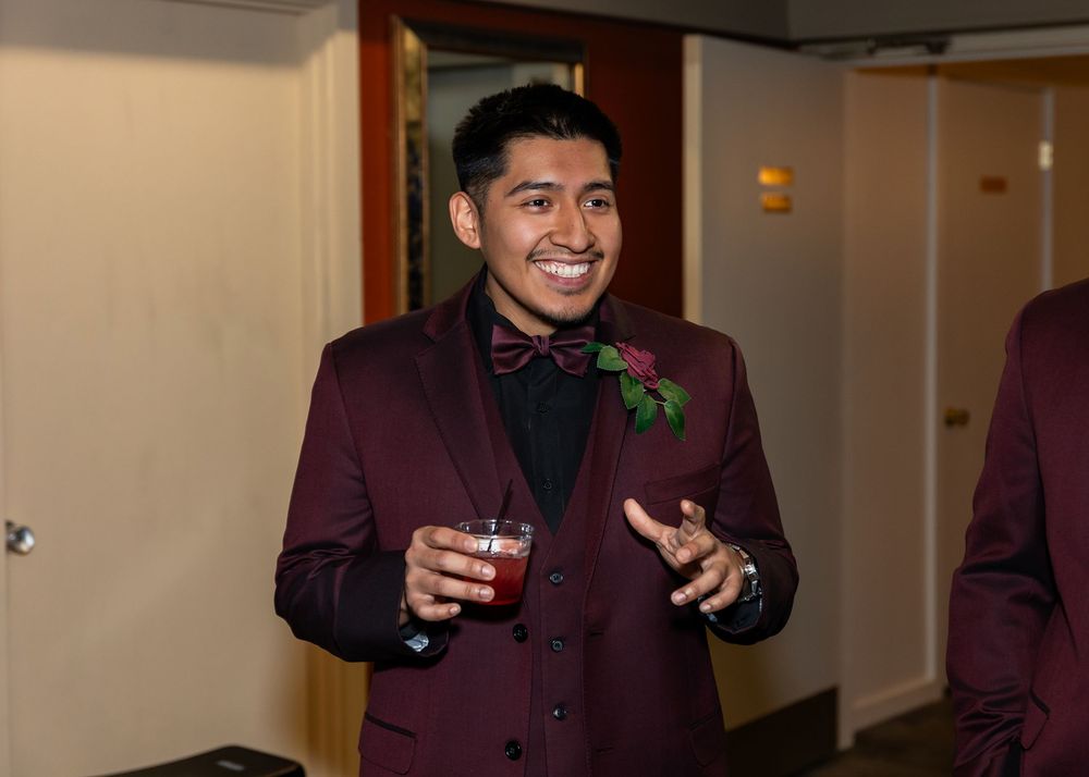 Young man in burgundy suit with bow tie and rose boutonniere holding a drink and smiling at a formal event.