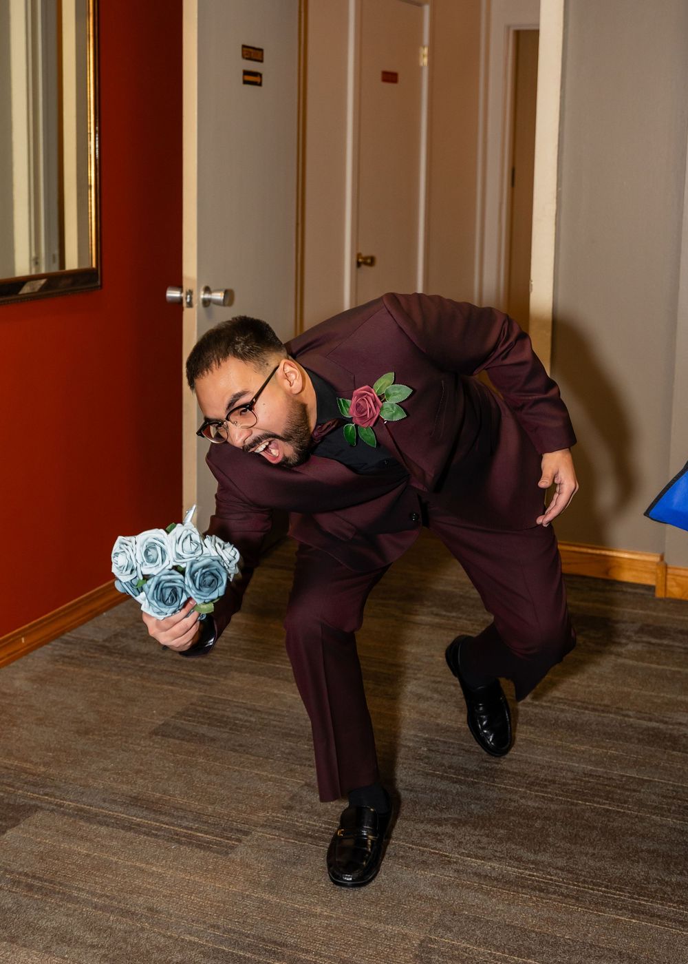 Man in burgundy suit holding blue roses bouquet strikes fun pose in hallway at wedding event.