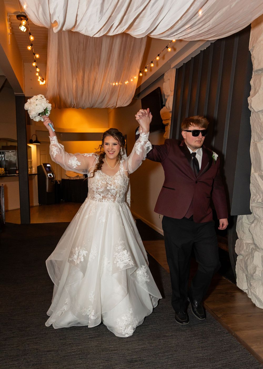 Bride and groom celebrate their grand entrance at wedding reception, raising hands triumphantly under draped fabric ceiling.