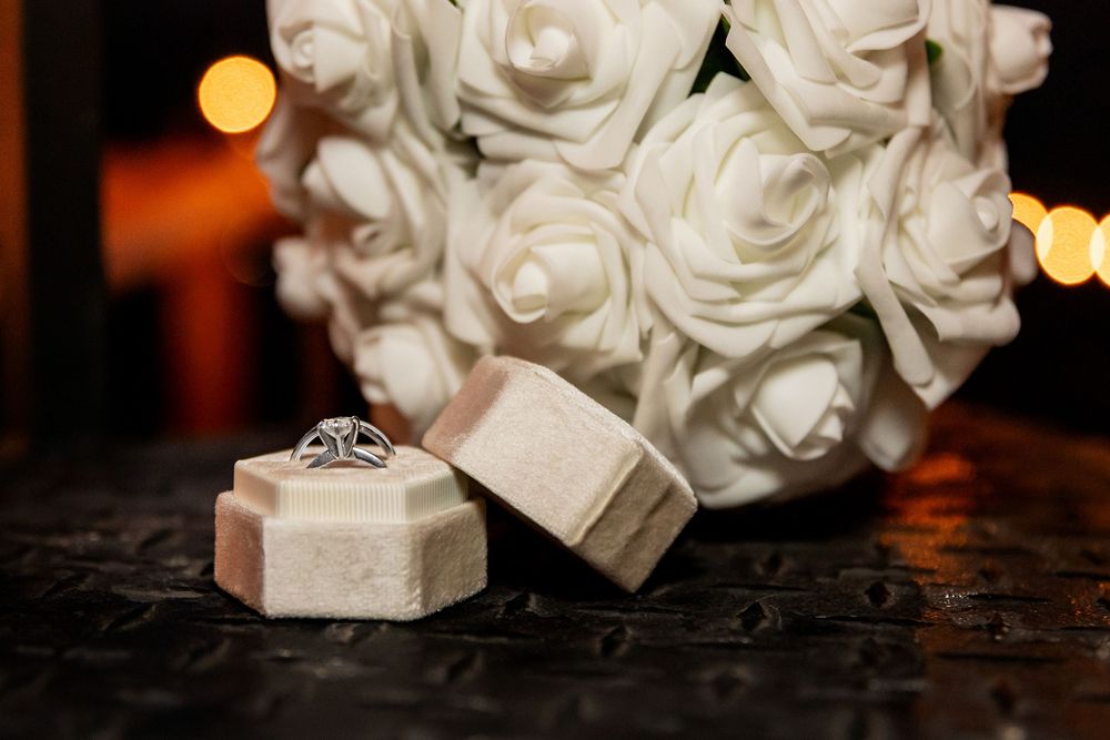 Diamond engagement ring in a beige velvet box beside a white rose bouquet with warm bokeh lights in the background.