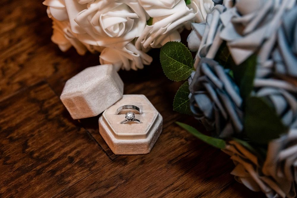 Close-up of wedding rings in a hexagonal velvet box surrounded by white and blue roses on a wooden surface.