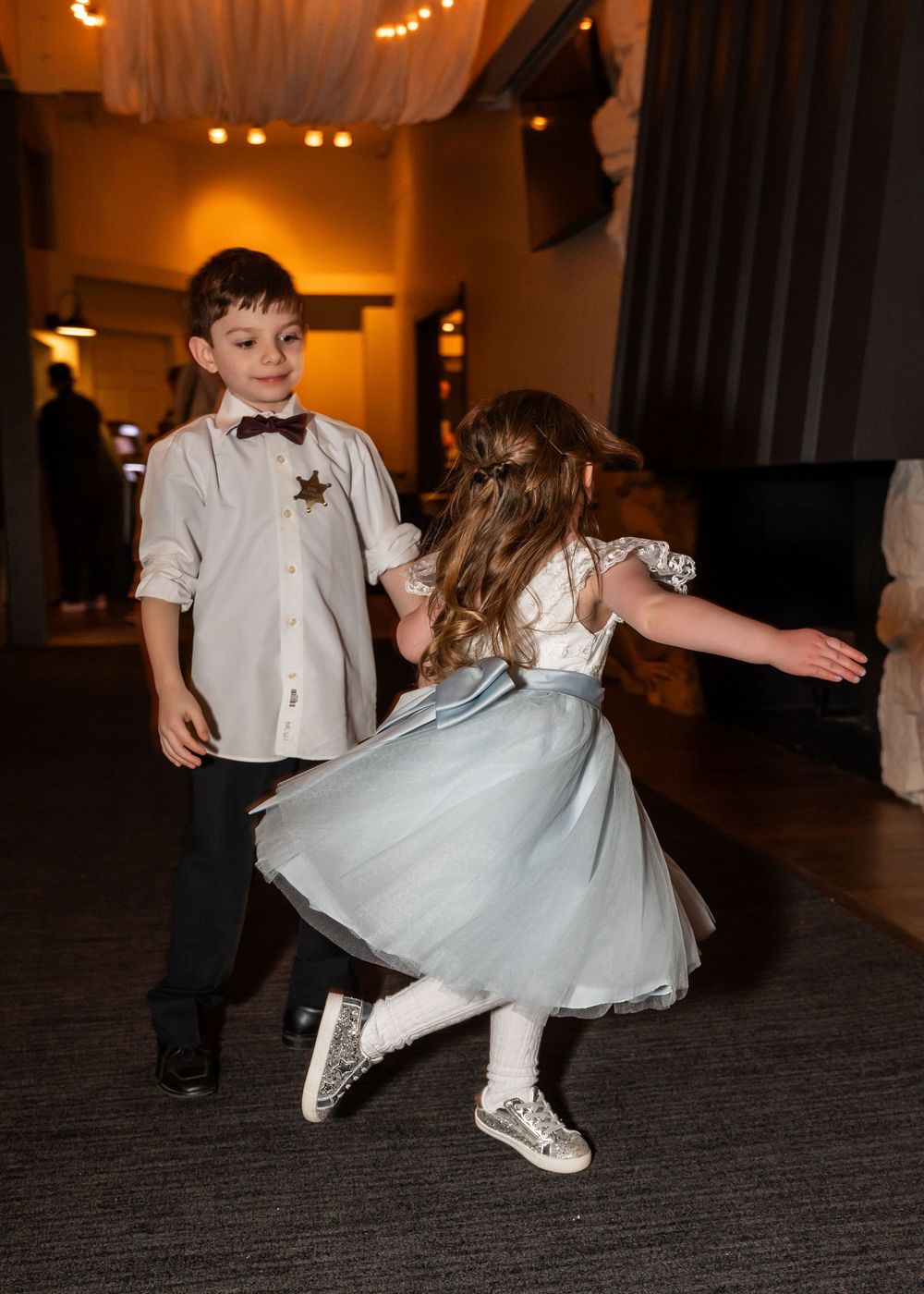 Two children dancing joyfully at a wedding reception, boy in bow tie and girl in blue tulle dress.
