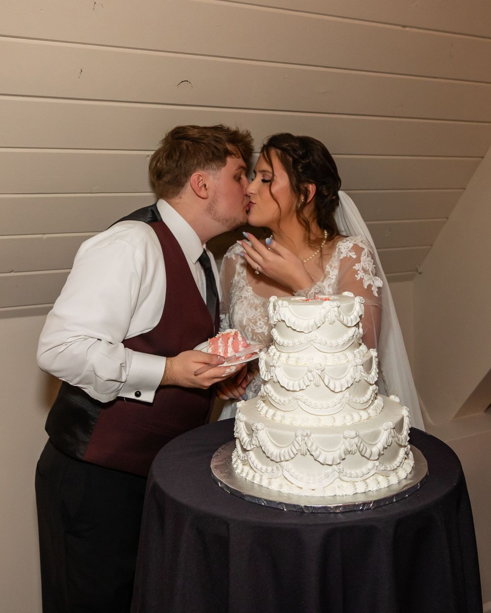 Bride and groom share a kiss while cutting their elegant four-tier white wedding cake at their reception.