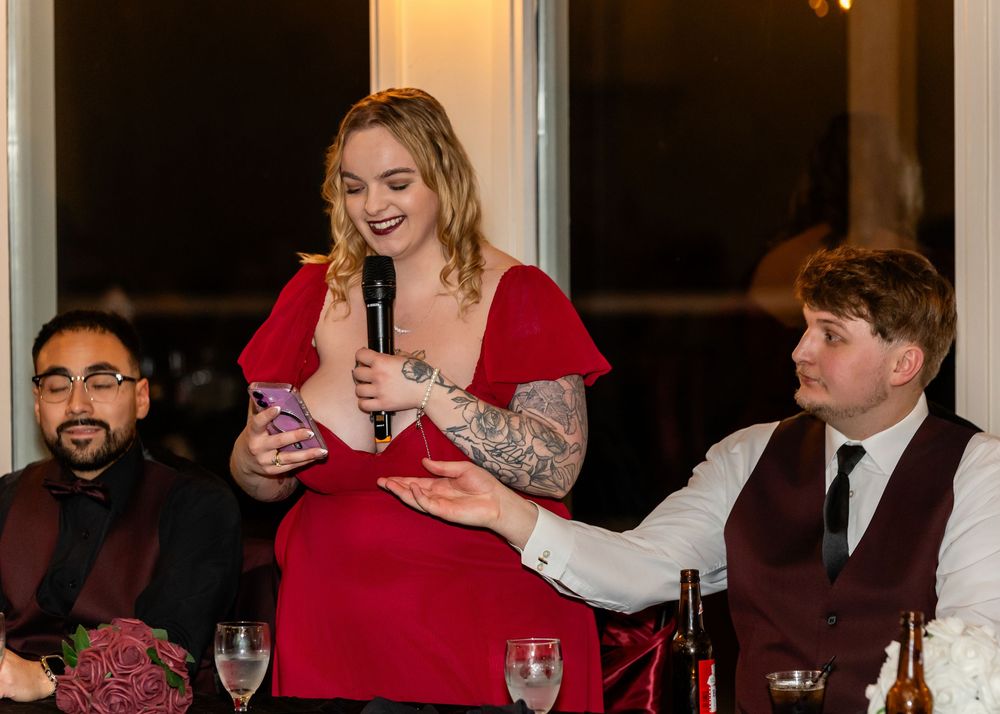 Woman in red dress giving a wedding toast with microphone while guests listen at the reception dinner table.