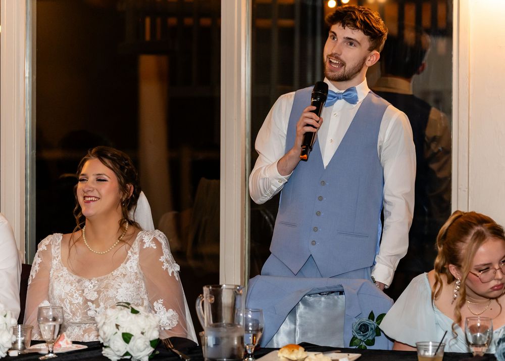 Groom in blue vest giving a speech at wedding reception while bride in lace dress smiles beside him.