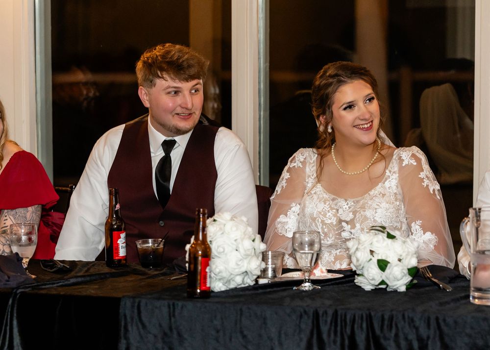 Bride and groom smiling at wedding reception head table with white floral centerpiece and candles.