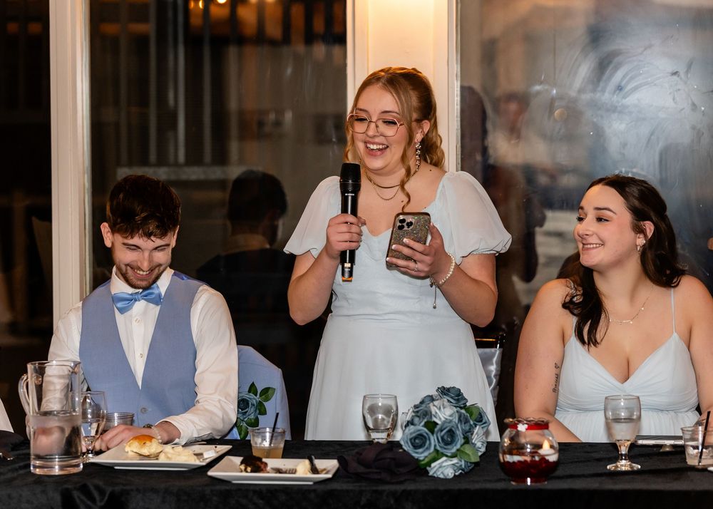 Wedding guest delivers heartfelt speech with microphone at reception dinner table with bride and groom seated nearby.