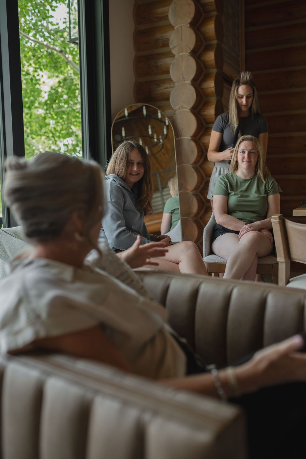 People sit and relax in a rustic log cabin lounge area with large windows and comfortable brown leather seating.