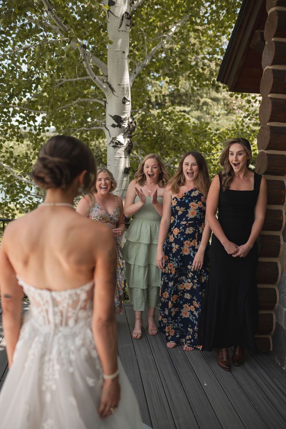 Bride in white dress reveals her gown to bridesmaids in floral and mint dresses on a covered wooden deck.