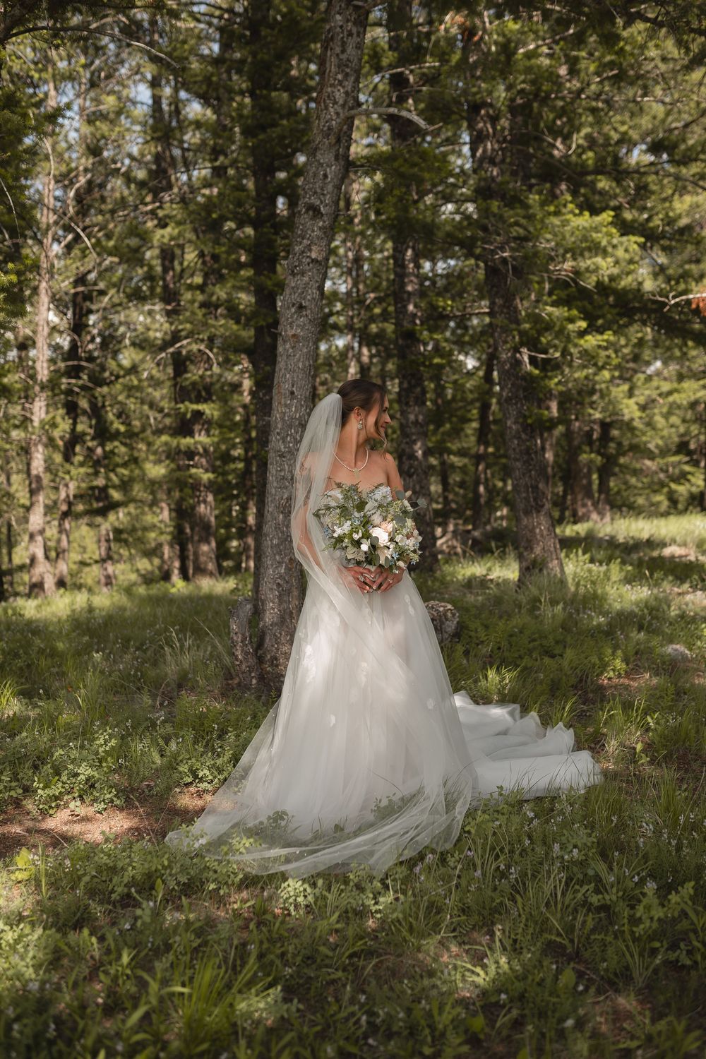A bride in a flowing white wedding gown stands gracefully among tall pine trees in a serene forest setting.
