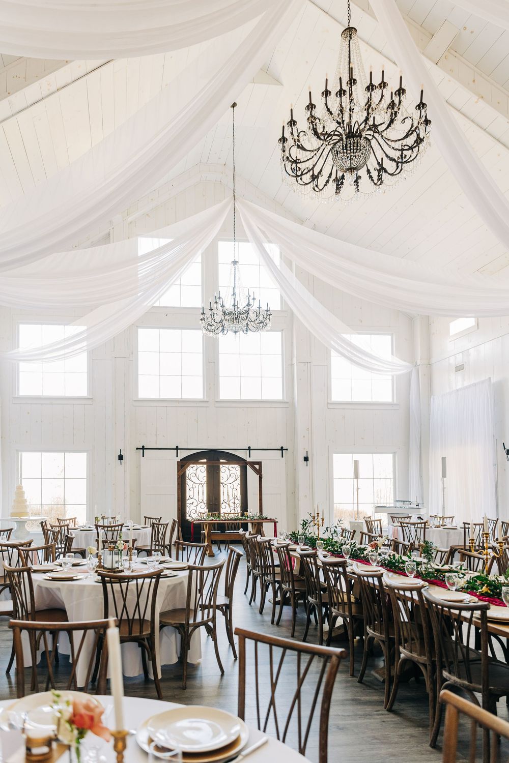Elegant white wedding reception venue with draped fabric ceilings, chandeliers, and long wooden dining tables.