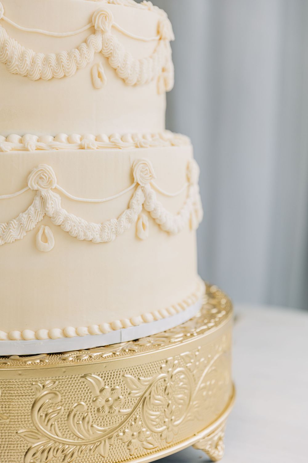 Elegant white wedding cake with delicate piping details sits on ornate gold decorative stand with intricate scroll patterns.