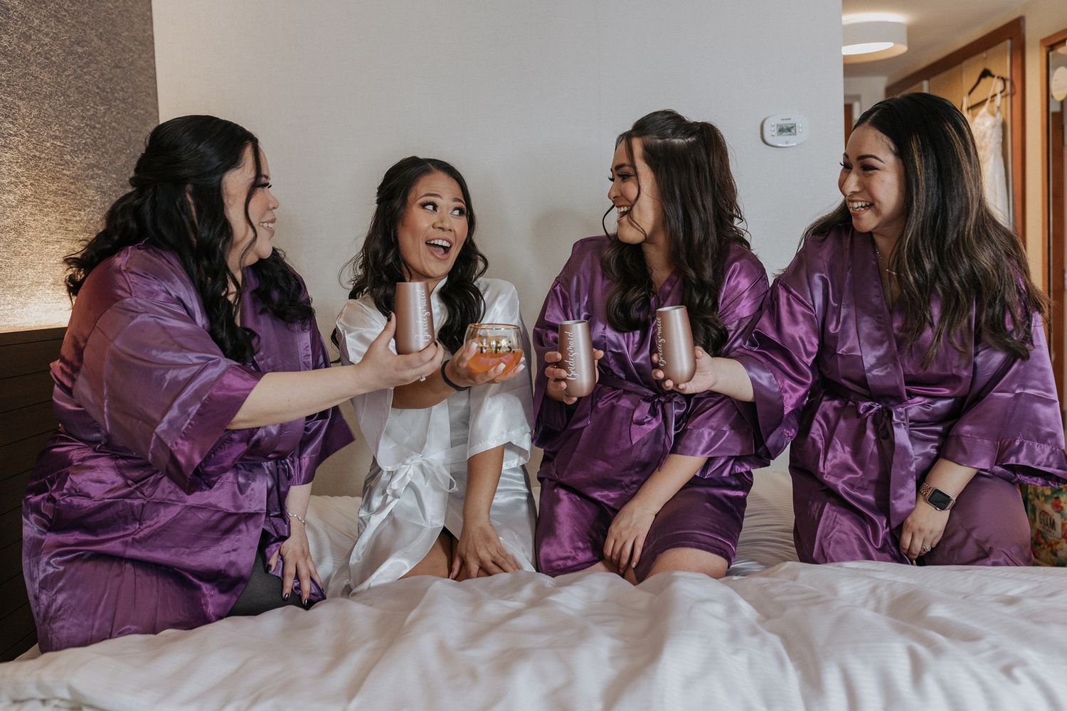 Bride and bridesmaids in matching purple satin robes sits on a bed laughing and enjoying a celebratory moment together.