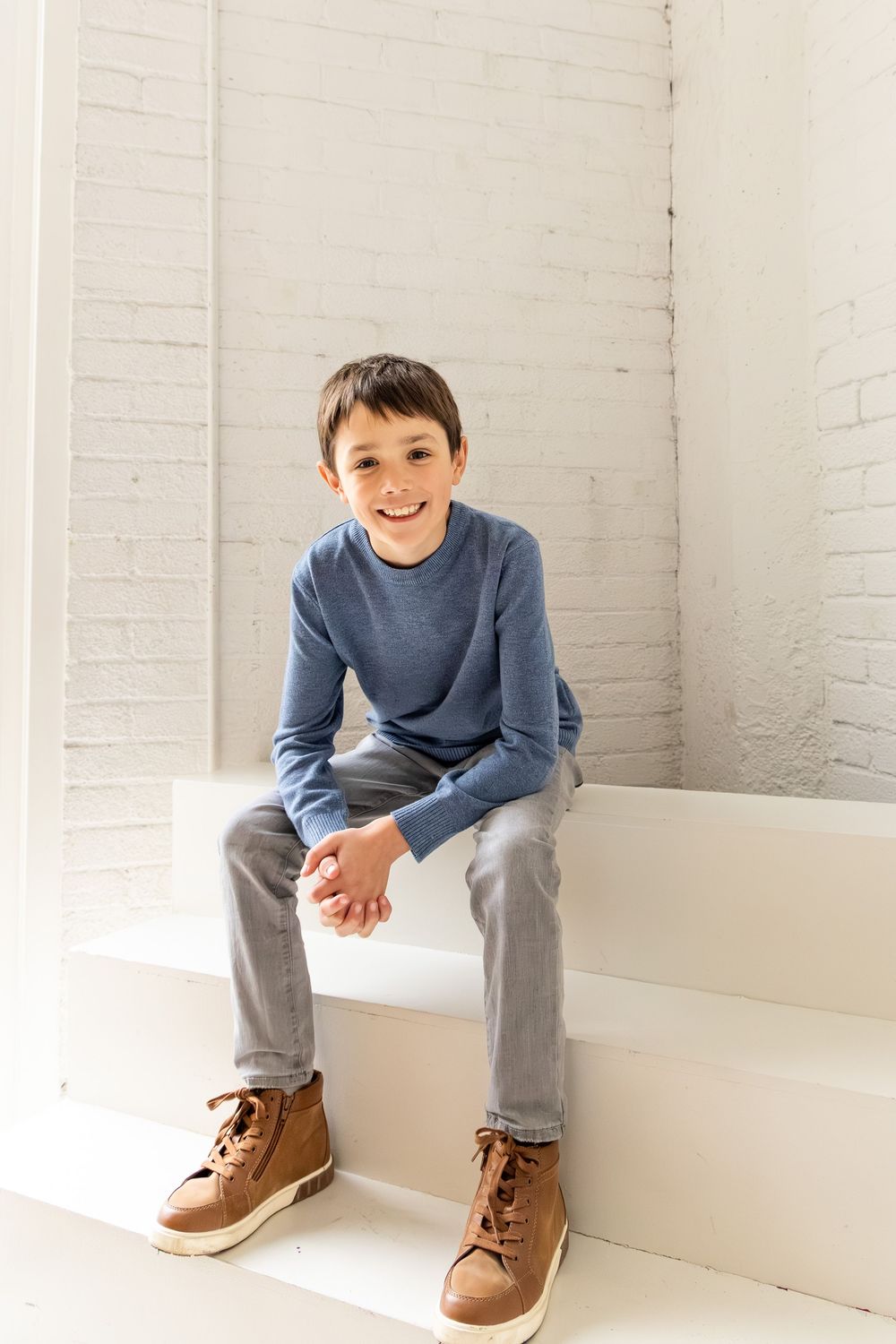 A figure in a blue sweater and gray pants sits casually on a white platform against a white brick wall.