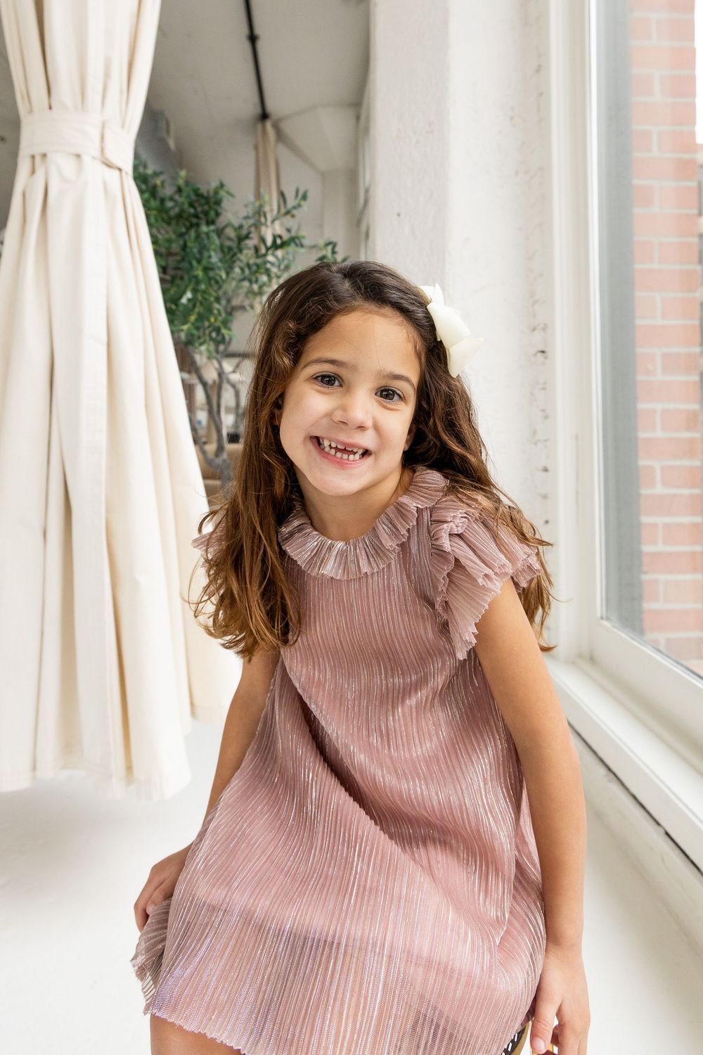 A young child in a pink ruffled top smiles while playing in front of white curtains and windows.