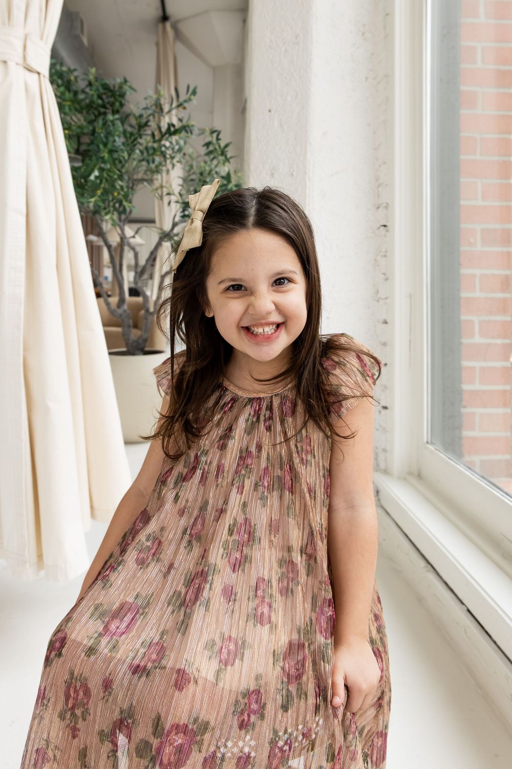 A child wearing a floral printed dress poses playfully near a bright window with white curtains.