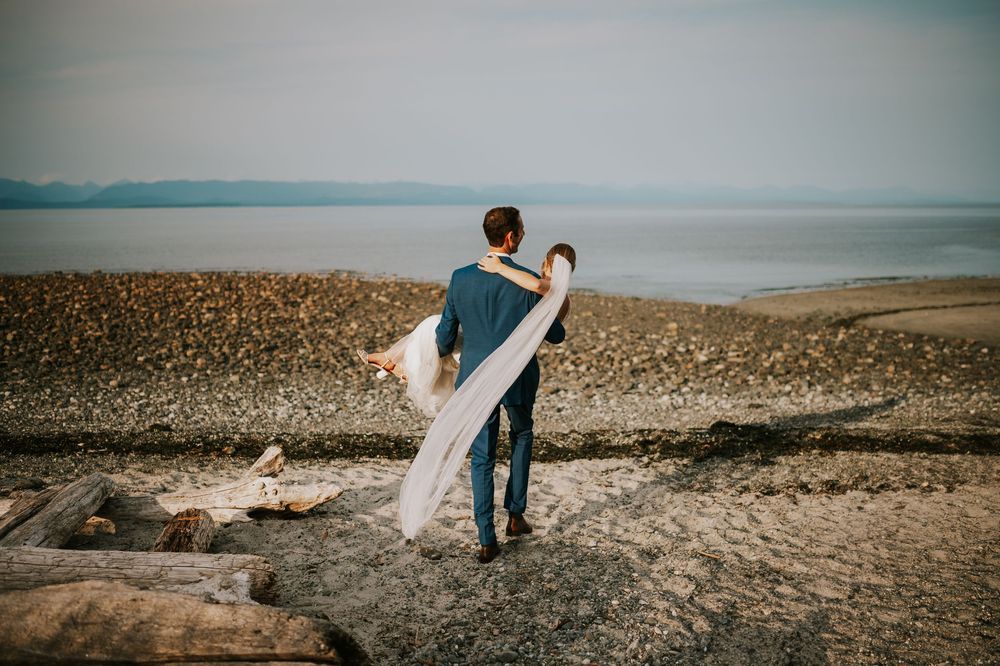 Wedding photography of couple on Williams Beach, groom carrying bride during golden hour with ocean, sand and mountains in background
