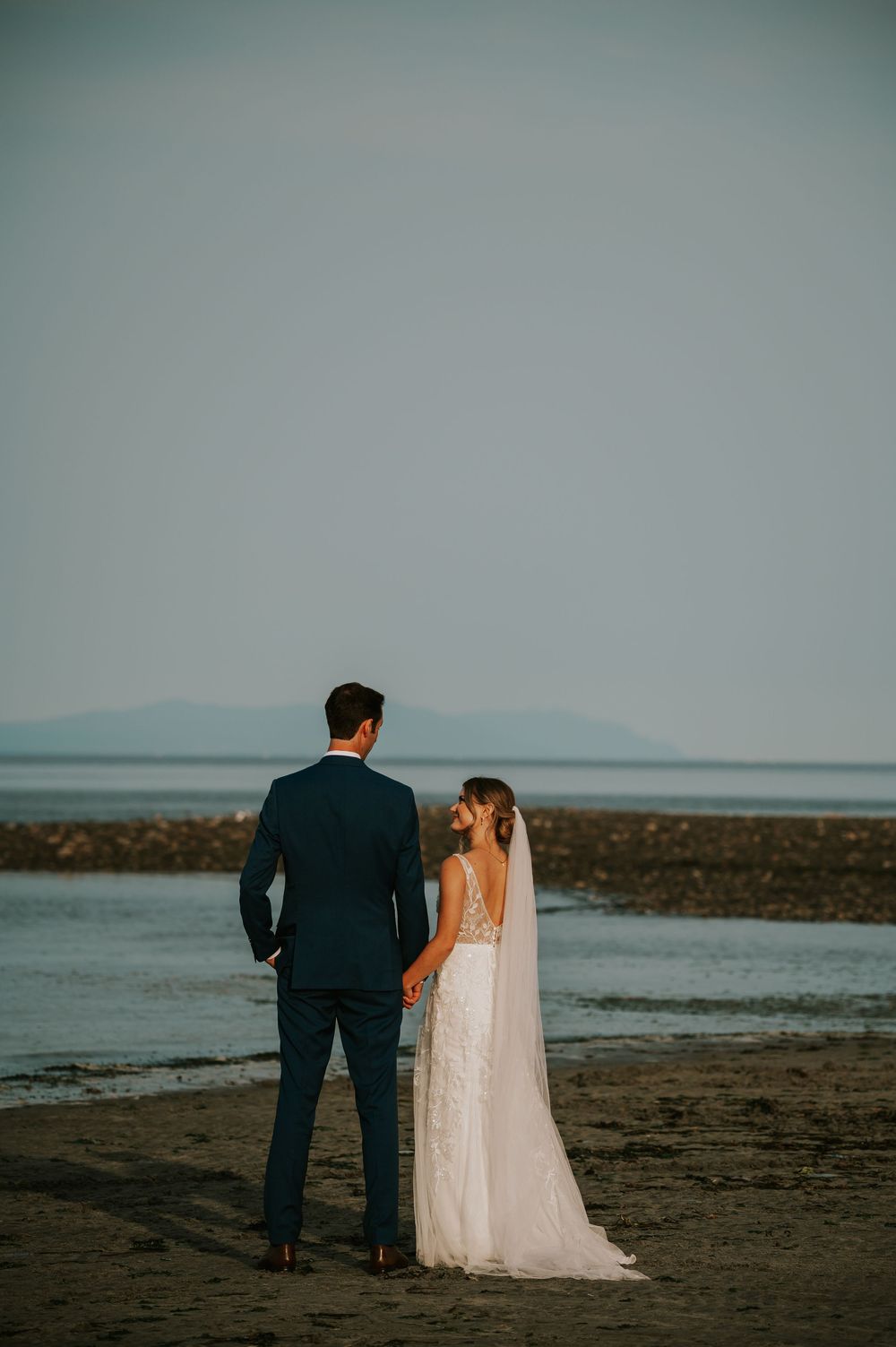 Wedding photography of couple on Williams Beach, groom and bride facing ocean during golden hour with ocean, and mountains in background
