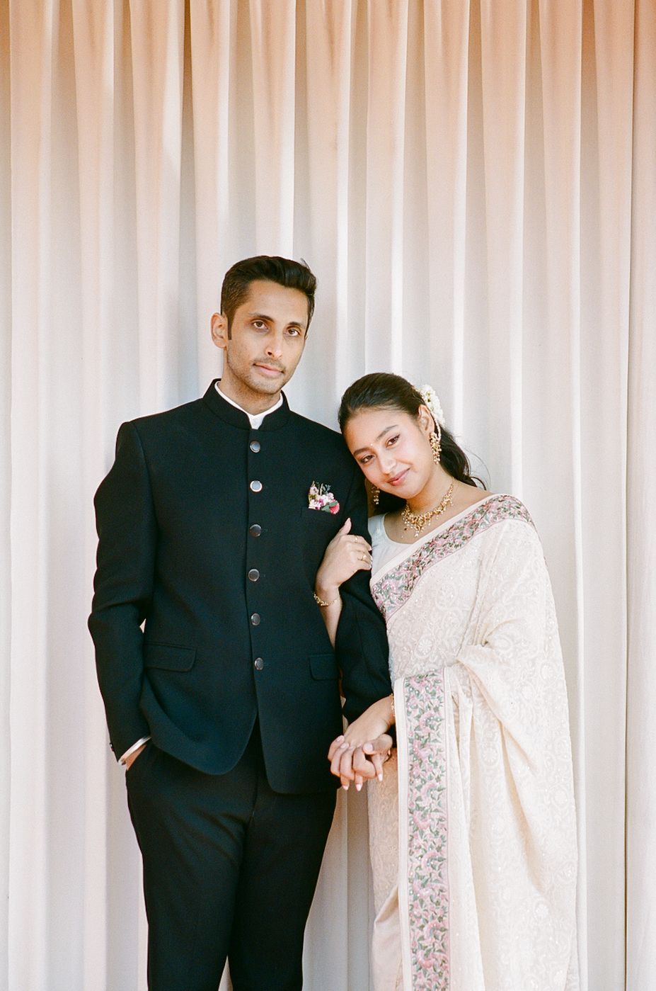 A couple poses together at a formal event, with one wearing a black suit and the other in an elegant white sari.