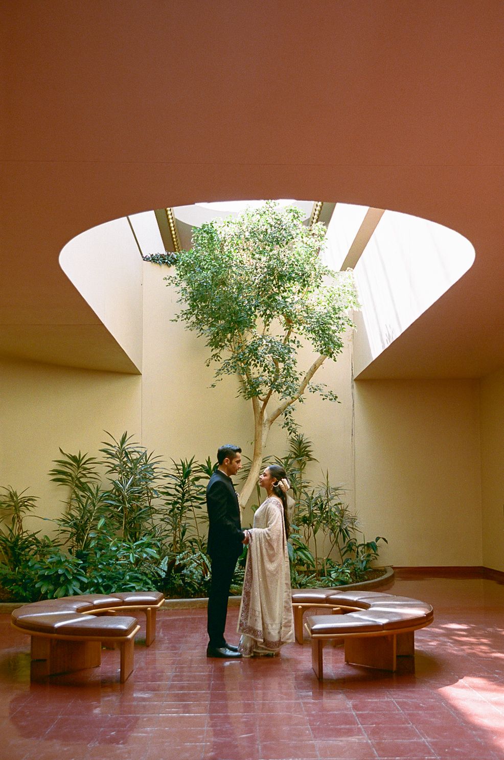 Two people stand together in a serene indoor courtyard with a skylight and potted plants on terra cotta floors.