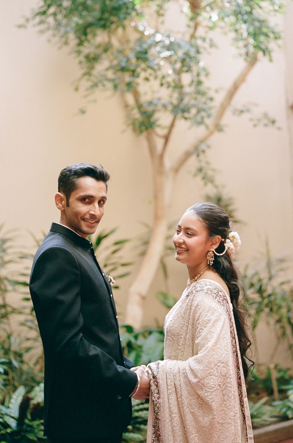 A couple in formal attire, with the person in black and person in beige, share a tender moment in a garden setting.
